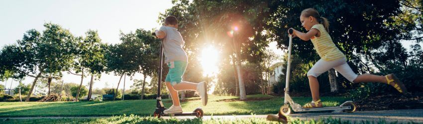 Children riding scooters through a sunlit park.